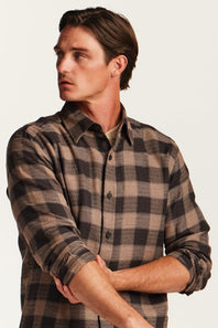 A man wearing a brown and beige plaid button-down flannel shirt, shown from the chest up against a plain off-white background.