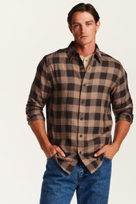 A man wearing a brown and beige plaid button-down shirt over a beige undershirt, paired with blue jeans, standing against a plain off-white background.