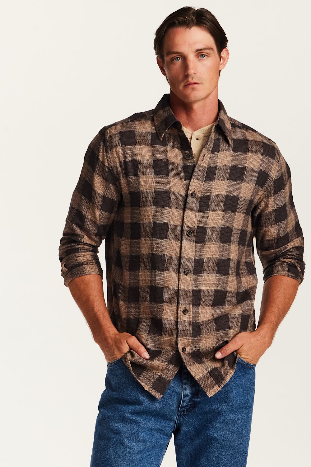 A man wearing a brown and beige plaid button-down shirt over a beige undershirt, paired with blue jeans, standing against a plain off-white background.