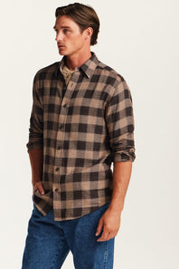 A man wearing a brown and beige buffalo plaid button-down flannel shirt with blue jeans, standing against a plain off-white background.
