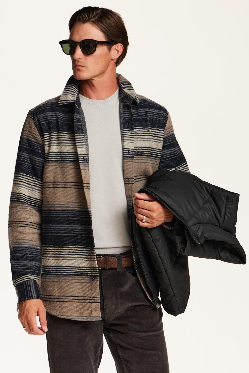 A man wearing a navy, beige, and brown striped button-up shirt jacket over a light gray t-shirt, holding a black puffer jacket, set against a plain off-white background.