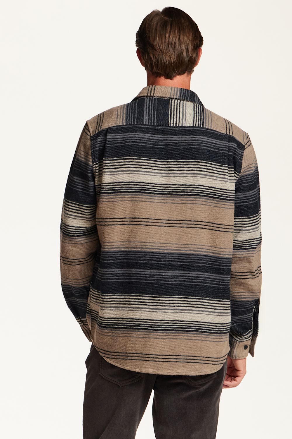 A man viewed from behind wearing a long-sleeve striped button-up shirt with horizontal bands of beige, brown, and dark navy, paired with dark gray pants, against a plain off-white background.