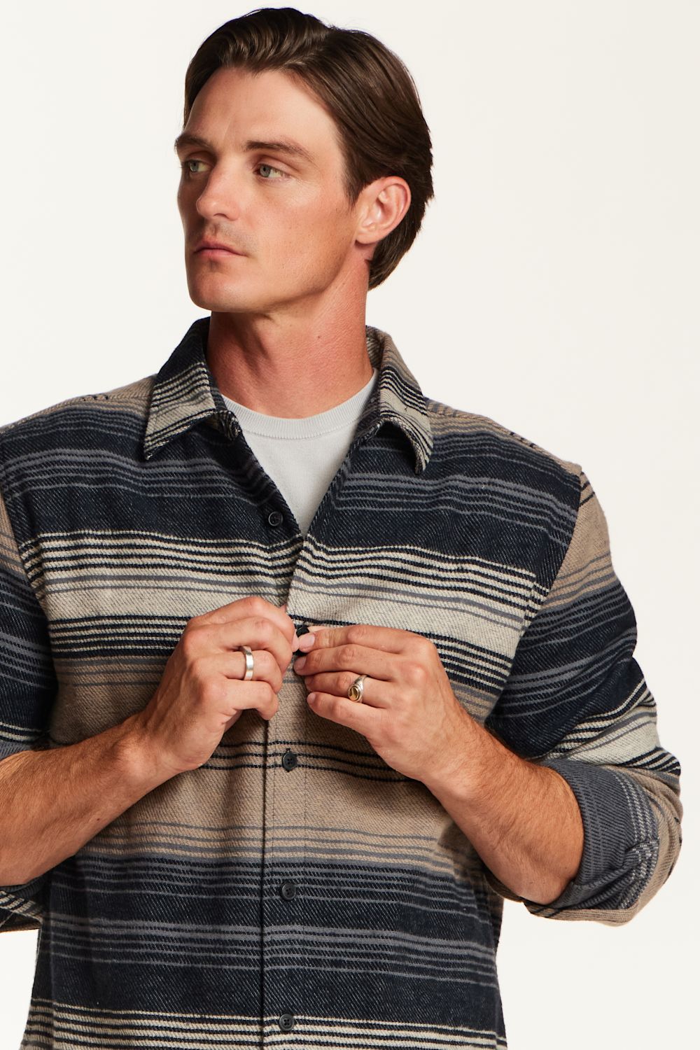 A man wearing a striped button-up shirt with navy, gray, and beige horizontal stripes over a light gray t-shirt, against a plain off-white background.