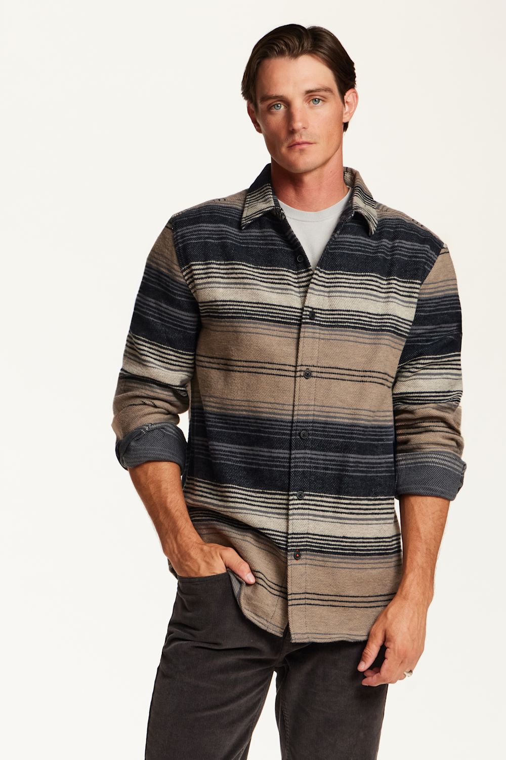 A man wearing a long-sleeve button-up shirt with horizontal stripes in navy, beige, and gray, layered over a light gray t-shirt and dark gray pants, against a plain white background.
