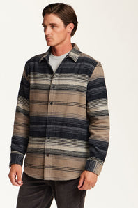 A man wearing a long-sleeve button-up shirt with horizontal stripes in navy blue, beige, and gray, layered over a light gray t-shirt and dark gray pants, photographed against a plain off-white background.