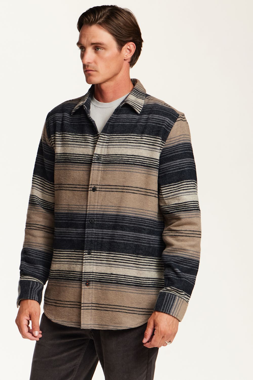 A man wearing a long-sleeve button-up shirt with horizontal stripes in navy blue, beige, and gray, layered over a light gray t-shirt and dark gray pants, photographed against a plain off-white background.