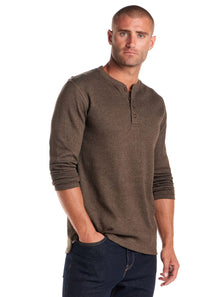 A man wearing a brown long-sleeve henley shirt and dark blue jeans, standing against a plain white background.