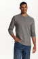 A man wearing a heather gray long-sleeve henley shirt with three buttons on the collar, paired with blue jeans, standing against a plain white studio background.