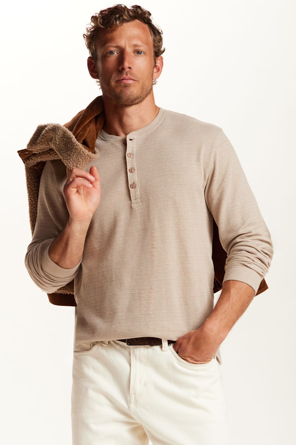 A man wearing a light beige long-sleeve henley shirt and white pants, with a brown jacket draped over his shoulder, photographed against a clean white studio background.