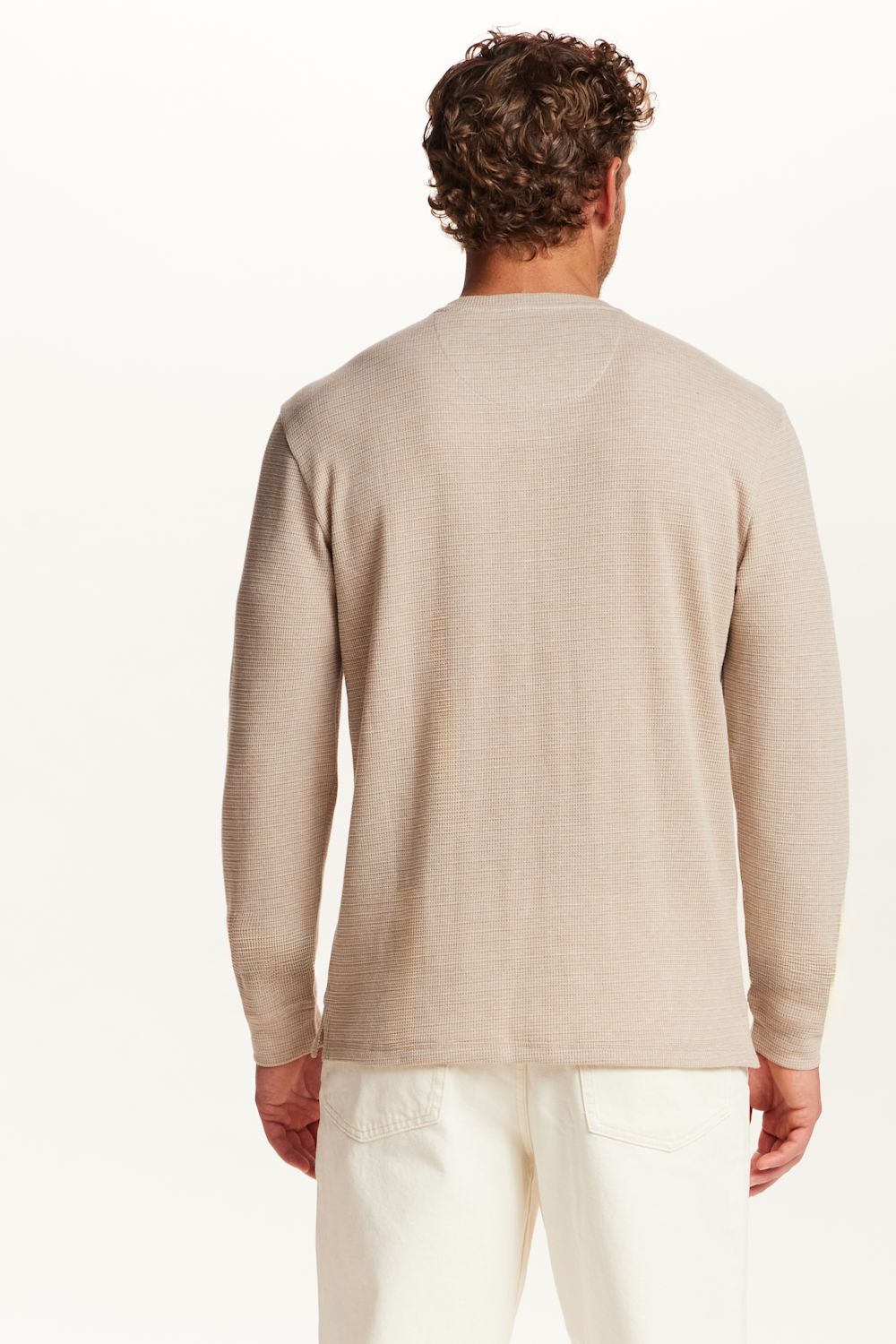 A man viewed from behind wearing a light beige long-sleeve knit sweater and off-white trousers, set against a plain white studio background.