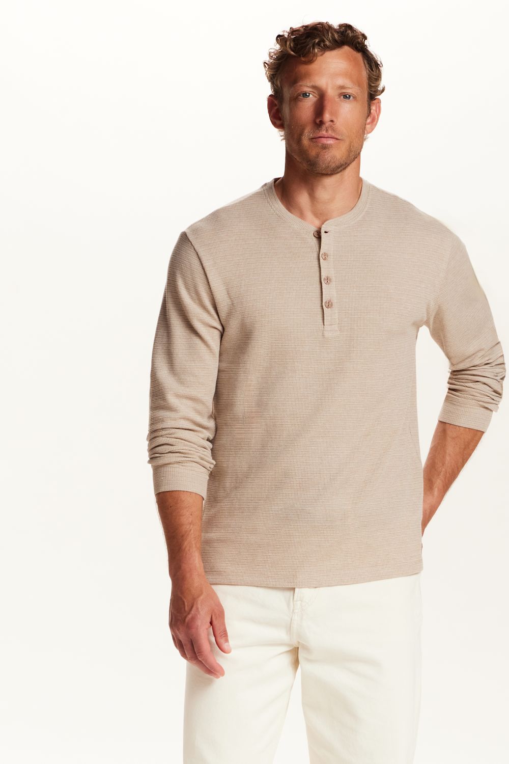 A man wearing a light beige long-sleeve henley shirt with white pants, standing against a plain white studio background.