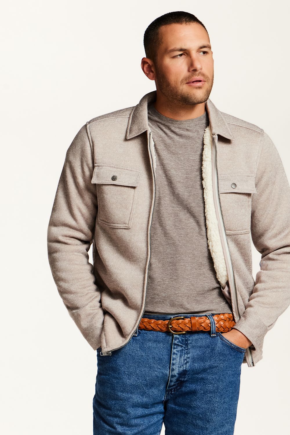 A man wearing a light beige sherpa-lined overshirt jacket over a gray t-shirt, blue jeans, and a brown woven belt, photographed against a plain white studio background.