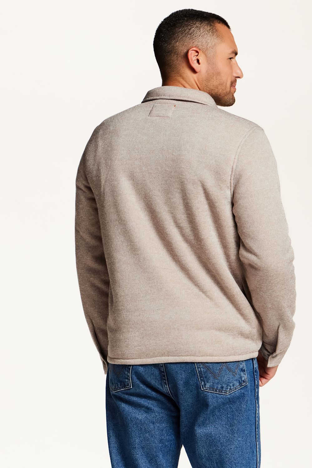 A man shown from the back wearing a light beige knit sweater and blue jeans against a plain white studio background.