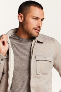A man wearing a light beige sherpa-lined zip-up jacket over a gray t-shirt, shown in a close-up profile shot against a plain off-white background.