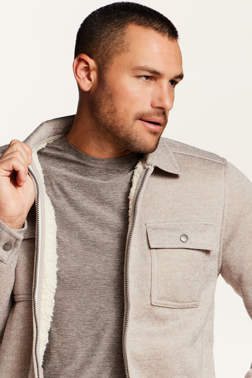 A man wearing a light beige sherpa-lined zip-up jacket over a gray t-shirt, shown in a close-up profile shot against a plain off-white background.