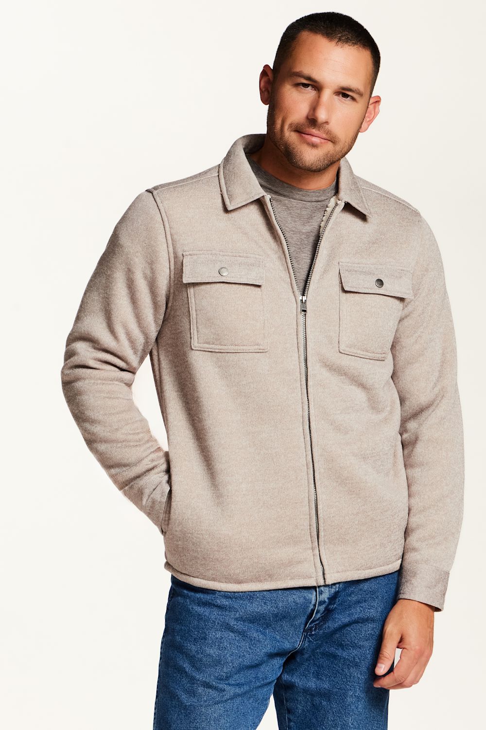 A man wearing a light beige zip-up fleece jacket over a gray t-shirt and blue jeans, photographed against a clean white studio background.
