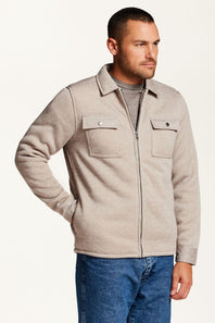 A man wearing a light beige sherpa-lined overshirt jacket over a gray t-shirt, blue jeans, and a brown woven belt, photographed against a plain white studio background.