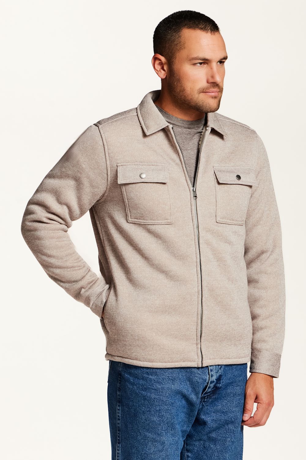 A man wearing a light beige sherpa-lined overshirt jacket over a gray t-shirt, blue jeans, and a brown woven belt, photographed against a plain white studio background.