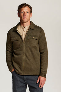 A man wearing an olive green zip-up fleece jacket over a beige collared shirt and dark blue jeans, standing against a light gray studio background.