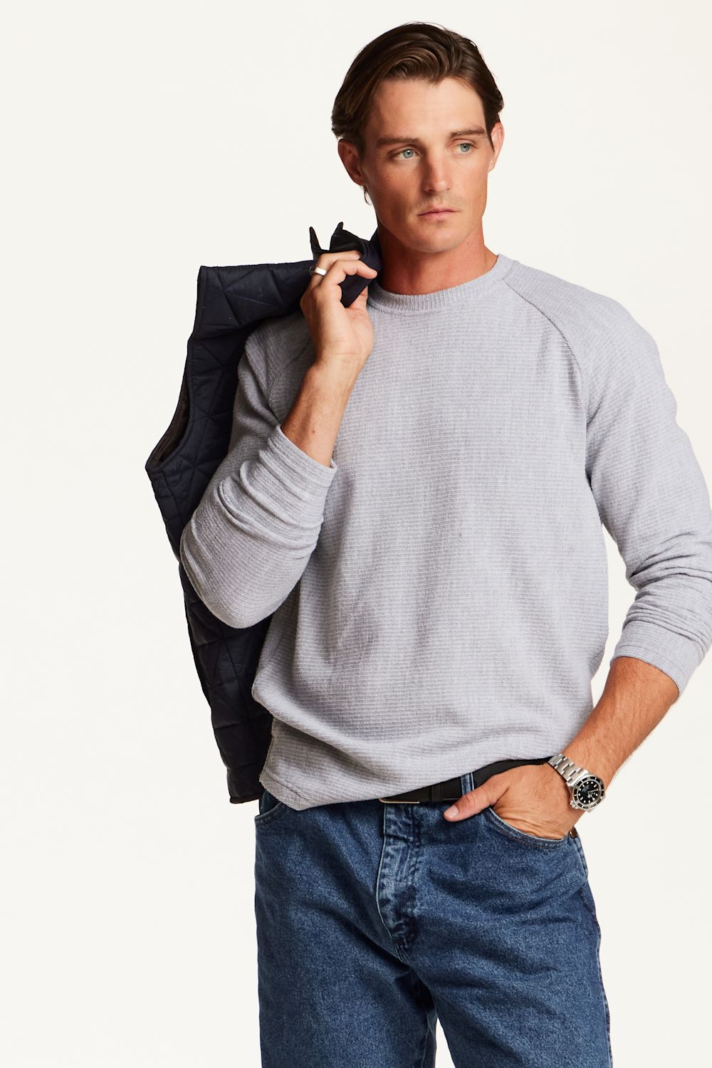 A man wearing a light gray long-sleeve knit crewneck sweater and blue jeans, holding a dark navy jacket over his shoulder against a plain off-white background.