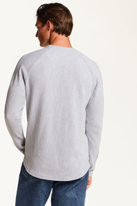 A man shown from the back wearing a light gray waffle-knit long-sleeve shirt and blue jeans, photographed against a plain off-white studio background.