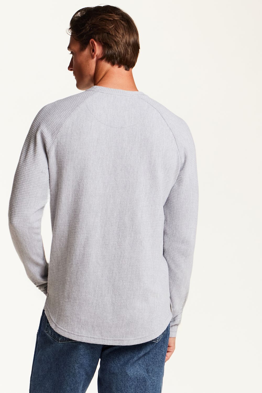 A man shown from the back wearing a light gray waffle-knit long-sleeve shirt and blue jeans, photographed against a plain off-white studio background.
