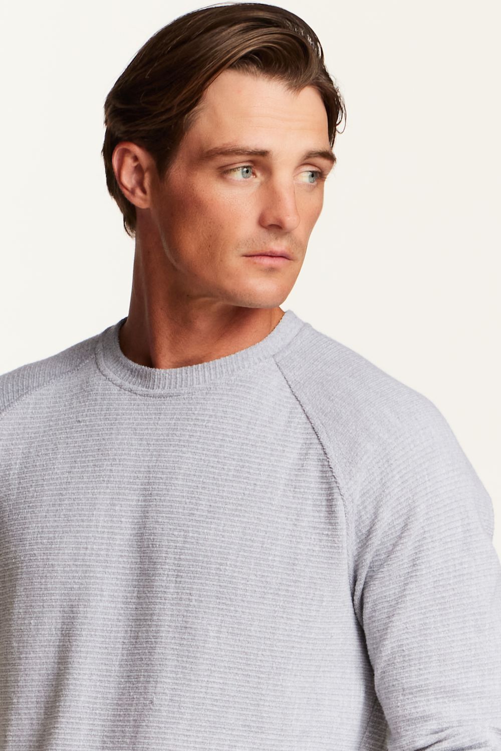 A close-up shot of a man wearing a light gray waffle-knit raglan crewneck sweater, shown from the chest up against a plain off-white background.