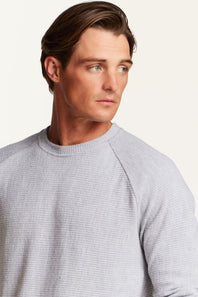 A close-up shot of a man wearing a light gray waffle-knit raglan crewneck sweater, shown from the chest up against a plain off-white background.