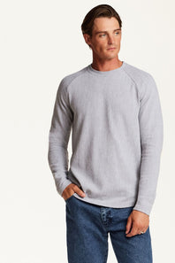 A man wearing a light gray waffle-knit long-sleeve raglan sweater and blue jeans, standing against a plain off-white studio background.