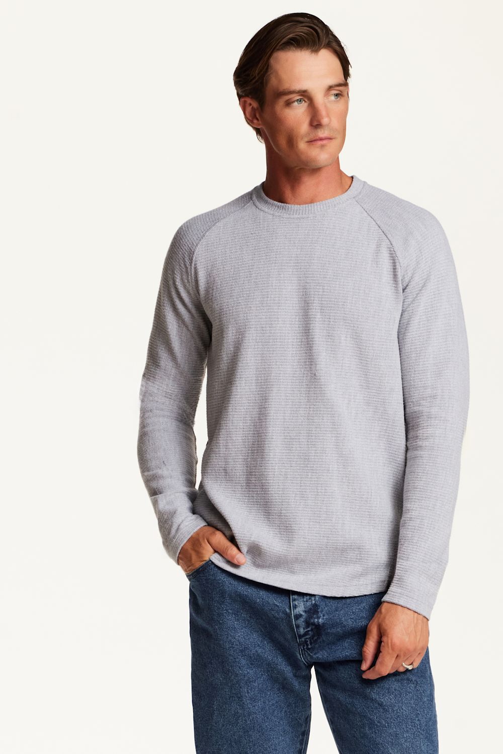 A man wearing a light gray waffle-knit long-sleeve raglan sweater and blue jeans, standing against a plain off-white studio background.