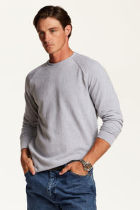 A man wearing a light gray waffle-knit long-sleeve raglan sweater and blue jeans, standing against a plain off-white studio background.