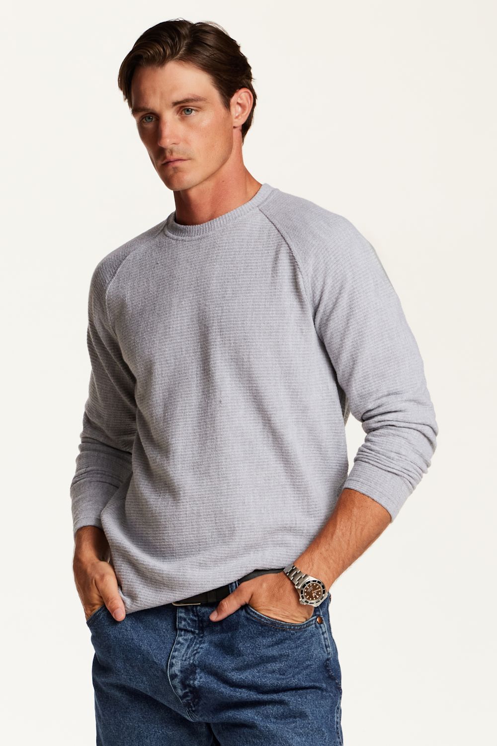 A man wearing a light gray waffle-knit long-sleeve raglan sweater and blue jeans, standing against a plain off-white studio background.