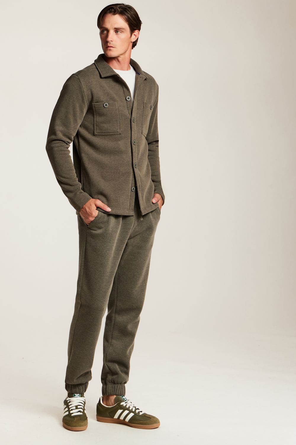 A man modeling a matching two-piece lounge set consisting of an olive green button-up shirt and jogger pants, paired with olive green and white sneakers, against a plain light gray studio background.