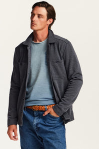 A man wearing a dark gray heathered button-up overshirt jacket over a light blue crewneck sweater, paired with blue jeans and a brown patterned belt, against a plain off-white studio background.
