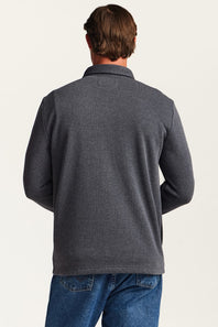 A man viewed from the back wearing a dark heather gray long-sleeve polo shirt and blue jeans against a plain off-white studio background.