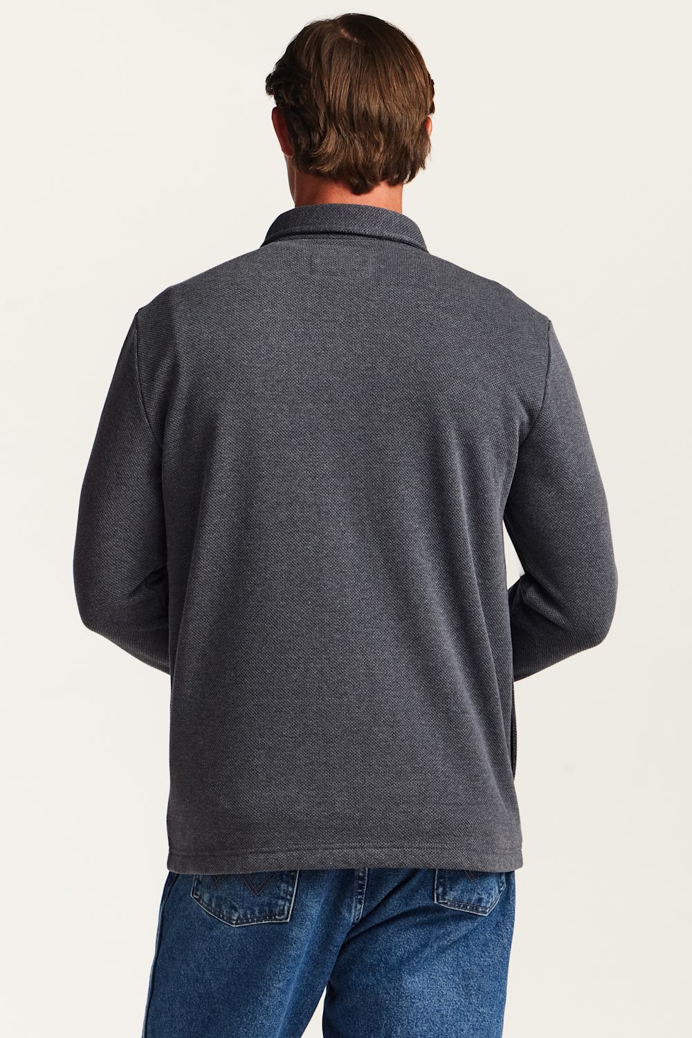 A man viewed from the back wearing a dark heather gray long-sleeve polo shirt and blue jeans against a plain off-white studio background.