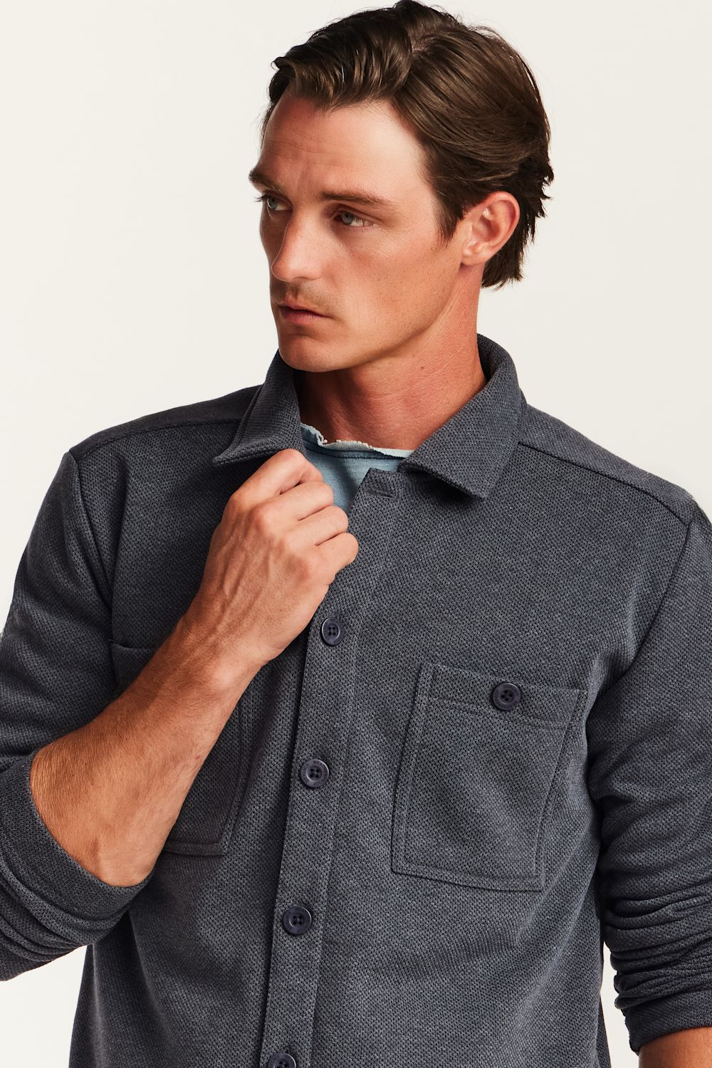 A man wearing a dark gray heathered button-up shirt with a chest pocket, shown in a close-up shot against a light off-white background.