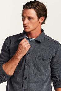 A man wearing a dark gray heathered button-up shirt with a chest pocket, shown in a close-up shot against a light off-white background.