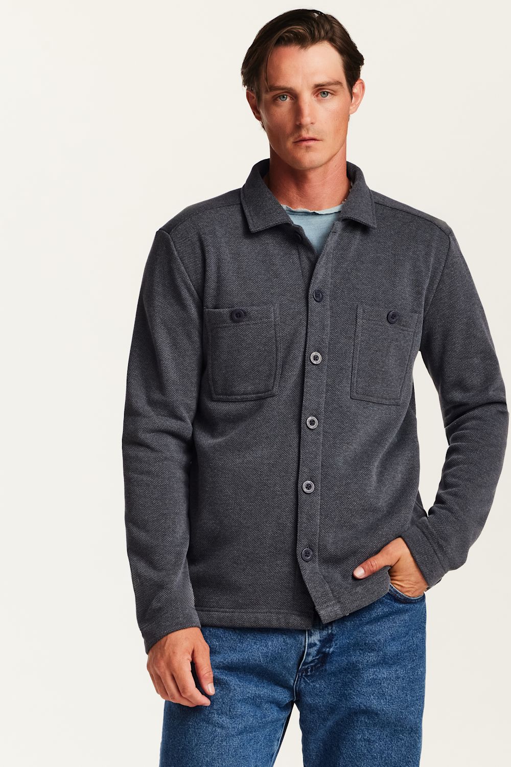 A man wearing a dark gray heathered button-up shirt jacket over a light blue t-shirt and blue jeans, standing against a plain off-white studio background.