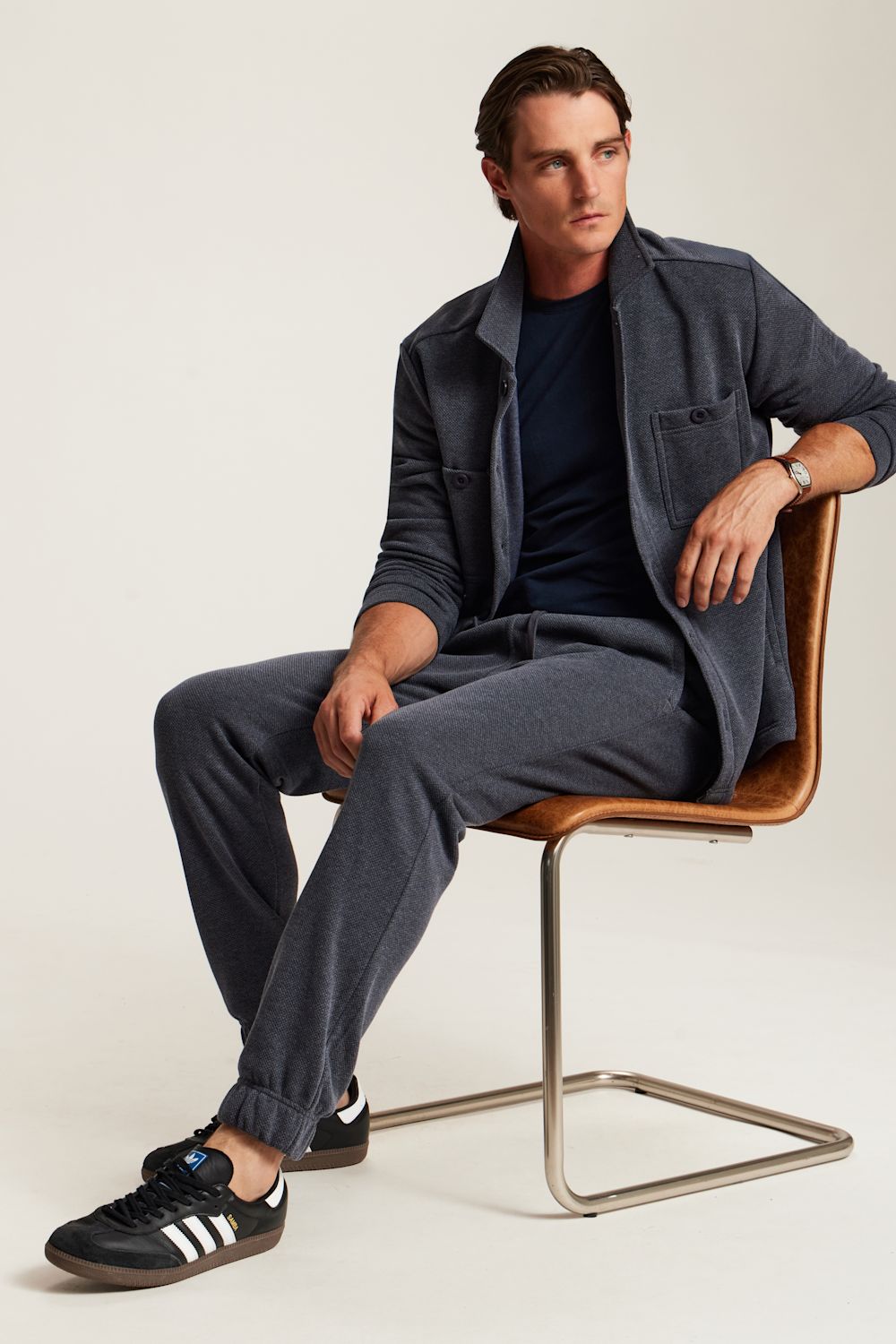 A man sits on a modern brown leather and metal chair wearing a dark gray overshirt, matching gray trousers, a navy t-shirt, and black Adidas sneakers against a light beige studio background.