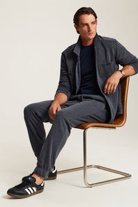 A man sits on a modern brown leather and metal chair wearing a dark gray overshirt, matching gray trousers, a navy t-shirt, and black Adidas sneakers against a light beige studio background.