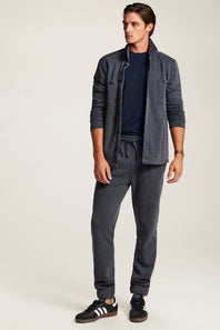 A man modeling a dark gray two-piece lounge set consisting of a lightweight zip-up jacket and matching jogger pants, styled with a navy t-shirt and black sneakers on a light beige studio background.