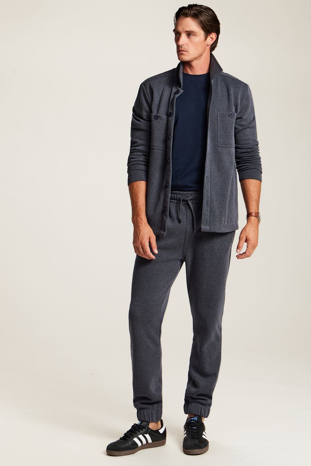 A man modeling a dark gray two-piece lounge set consisting of a lightweight zip-up jacket and matching jogger pants, styled with a navy t-shirt and black sneakers on a light beige studio background.