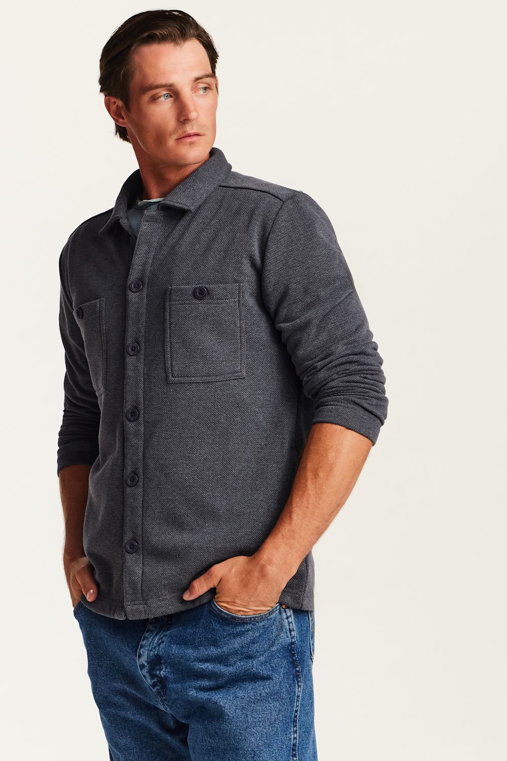 A man wearing a dark charcoal gray long-sleeve button-up shirt with a chest pocket, paired with blue jeans, photographed against a plain light background.