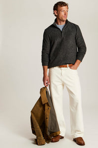 A man stands against a light gray studio background wearing a dark charcoal ribbed quarter-zip sweater over a light blue shirt, off-white trousers, and brown shoes. He holds a brown jacket in one hand.