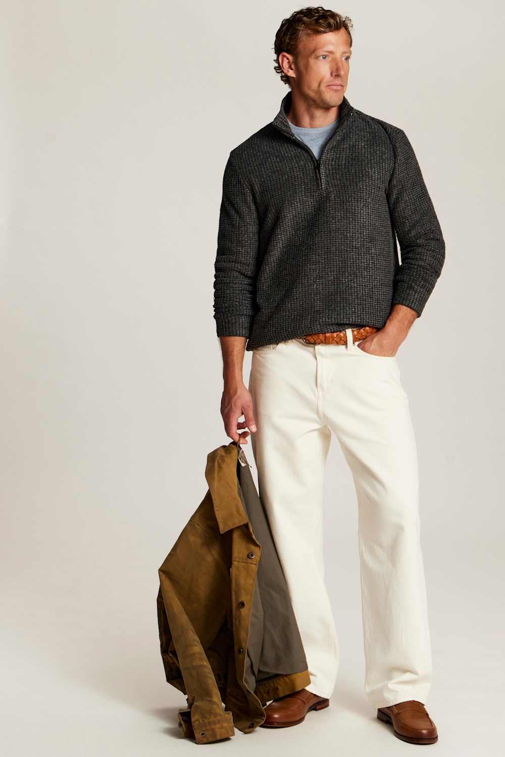 A man stands against a light gray studio background wearing a dark charcoal ribbed quarter-zip sweater over a light blue shirt, off-white trousers, and brown shoes. He holds a brown jacket in one hand.
