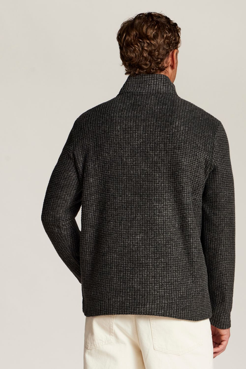A man viewed from the back wearing a dark charcoal gray textured knit sweater with a mock neck collar, paired with off-white trousers, against a light gray studio background.