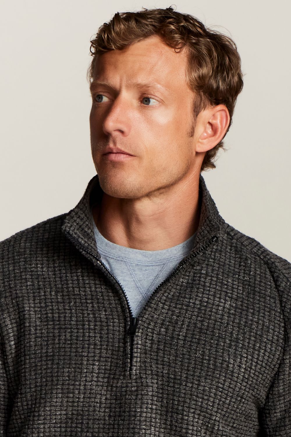 A man wearing a dark gray textured quarter-zip pullover sweater over a light blue t-shirt, photographed against a solid light gray background.