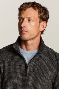 A man wearing a dark gray textured quarter-zip pullover sweater over a light blue t-shirt, photographed against a solid light gray background.