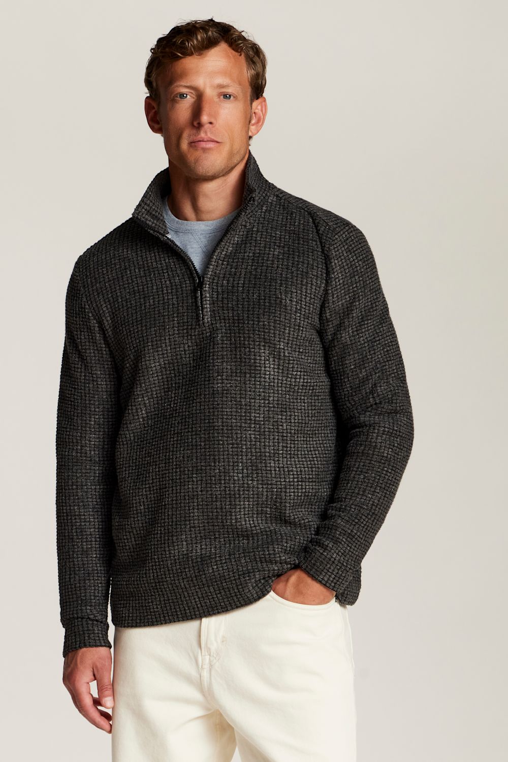 A man wearing a dark charcoal gray textured quarter-zip sweater over a light gray t-shirt and cream-colored pants, standing against a solid light gray background.